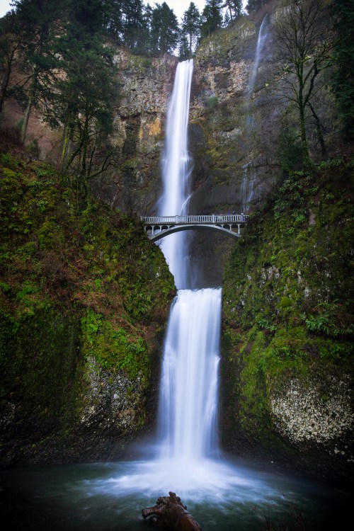 A stunning waterfall cascades down rocky cliffs, with a bridge spanning above, surrounded by lush greenery.