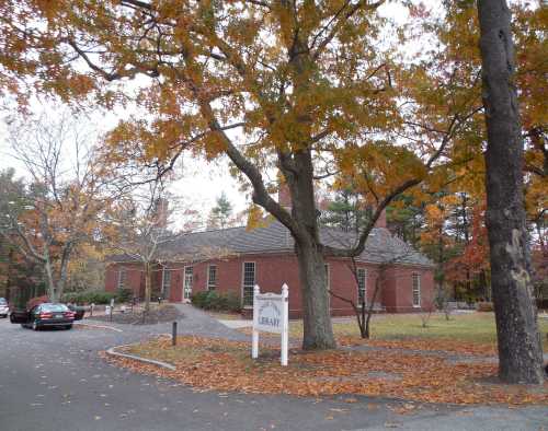 A brick library building surrounded by autumn trees with colorful leaves, parked cars in the foreground.