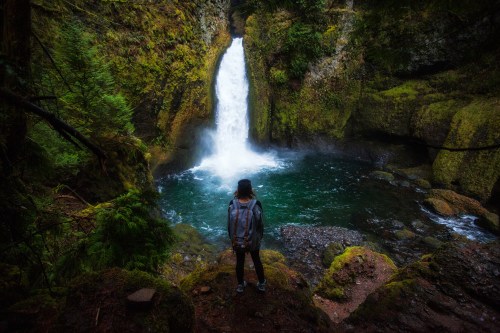A person stands near a waterfall surrounded by lush greenery and rocky terrain, gazing at the cascading water.