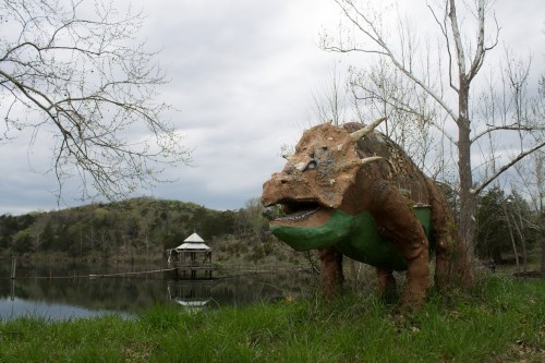 A large dinosaur sculpture stands by a pond, with a gazebo visible in the background under a cloudy sky.