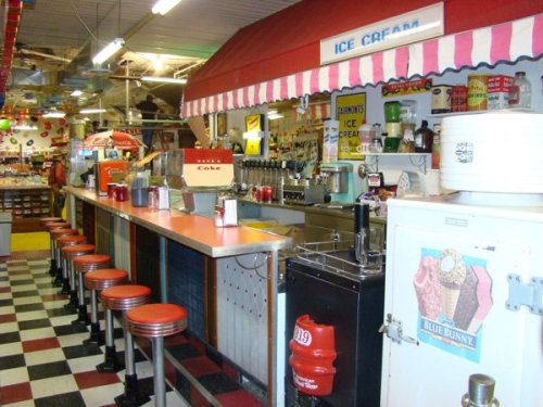 A retro ice cream shop featuring a long counter with red stools, colorful decor, and vintage ice cream machines.