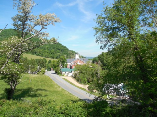 A scenic view of a small town with a road, green hills, and a building with a clock tower under a clear blue sky.