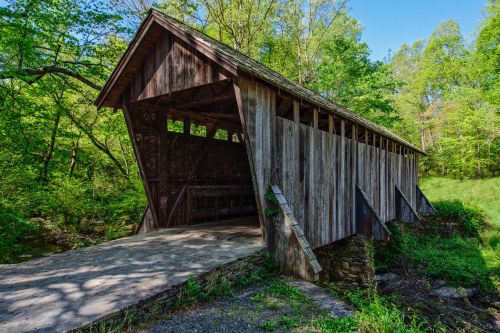 A rustic wooden covered bridge surrounded by lush green trees and a clear blue sky.