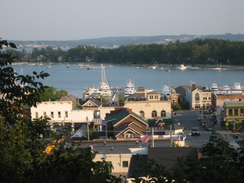 A scenic view of a coastal town with boats in the harbor, surrounded by trees and buildings under a clear sky.