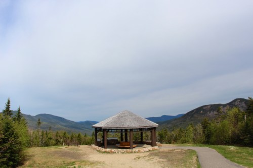 A wooden gazebo sits on a hillside, surrounded by green trees and mountains under a cloudy sky.