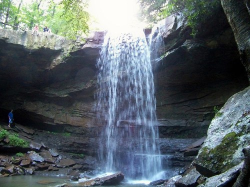 A waterfall cascades over rocky cliffs into a serene pool, surrounded by lush greenery and people observing from above.