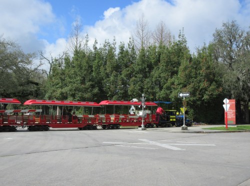 A red train with open-air cars is parked near a green hedge and a directional sign on a sunny day.