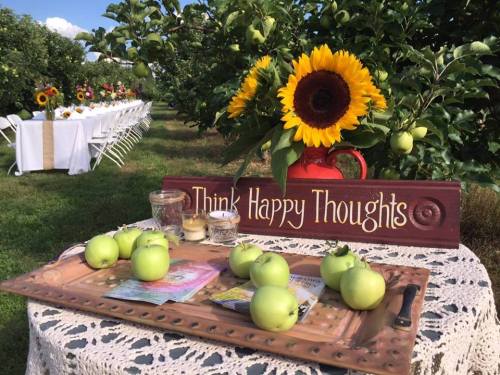 A rustic table with green apples, candles, and a sign reading "Think Happy Thoughts," surrounded by sunflowers and trees.
