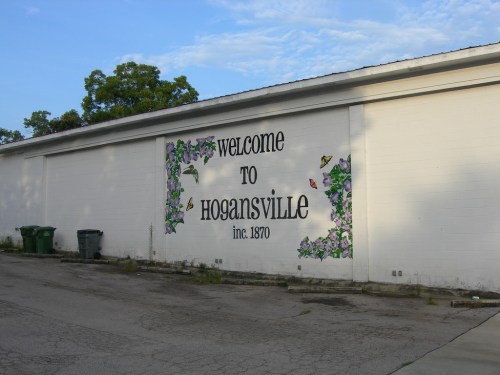Mural on a white wall reading "Welcome to Hogansville, Inc. 1870," surrounded by floral designs.