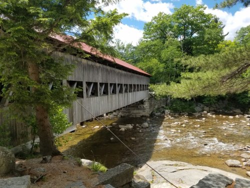 A wooden covered bridge spans a river, surrounded by lush greenery and rocky banks under a bright blue sky.