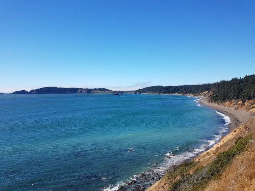 A scenic coastal view with clear blue water, sandy beach, and lush green hills under a bright blue sky.