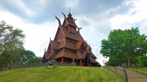 A tall, wooden church with intricate architecture, set on a grassy hill under a cloudy sky.
