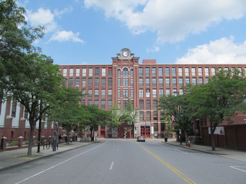 Historic brick building with a clock tower, flanked by trees, on a quiet street under a partly cloudy sky.