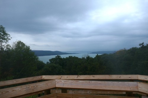 A cloudy view of a lake surrounded by trees, seen from a wooden deck.