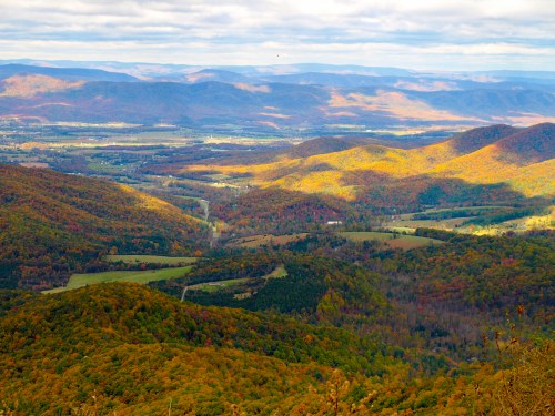 A panoramic view of rolling mountains and valleys, showcasing vibrant autumn foliage under a cloudy sky.