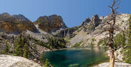 A serene mountain lake surrounded by rugged cliffs and trees under a clear blue sky.