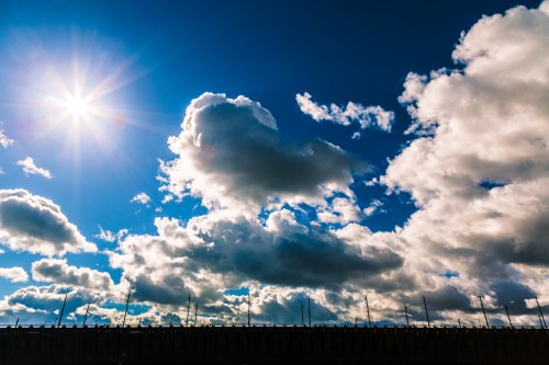 Bright sun shining over a blue sky filled with fluffy clouds and a silhouette of a fence in the foreground.