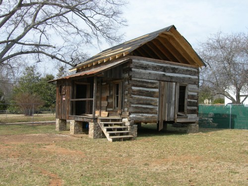 A rustic wooden cabin with a slanted roof, elevated on stone pillars, surrounded by grass and trees.