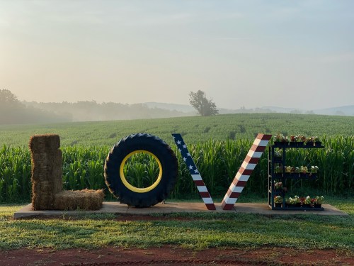 A large "LOVE" sign made of hay, a tire, and painted wood, set against a misty green field at sunrise.