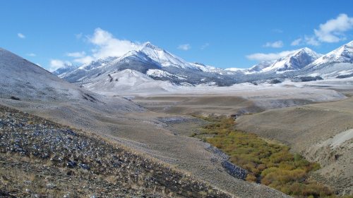 Snow-capped mountains rise over a valley with a winding river and autumn-colored trees under a clear blue sky.