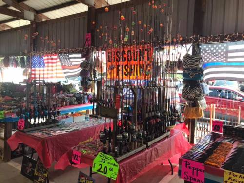 A market stall displaying fishing rods, tackle, and colorful hats, with American flags and discount signs in the background.