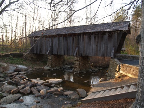 A wooden covered bridge spans a small stream, surrounded by trees and rocky terrain in a serene outdoor setting.