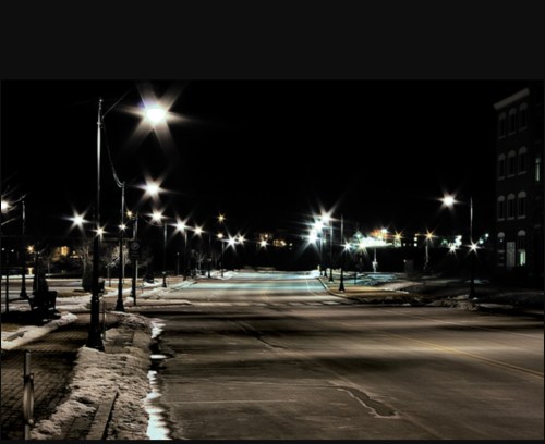 A quiet, empty street at night, illuminated by bright streetlights, with patches of snow on the ground.