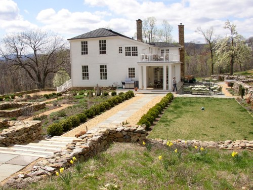 A white two-story house with a porch, surrounded by landscaped gardens and a stone pathway on a sunny day.