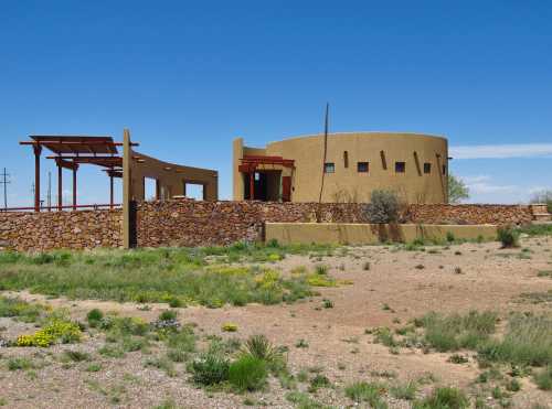 A round adobe building with a stone wall, surrounded by sparse vegetation under a clear blue sky.