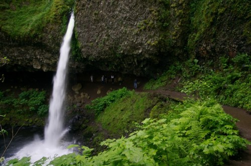 A waterfall cascades down rocky cliffs surrounded by lush greenery and a small group of people in the background.