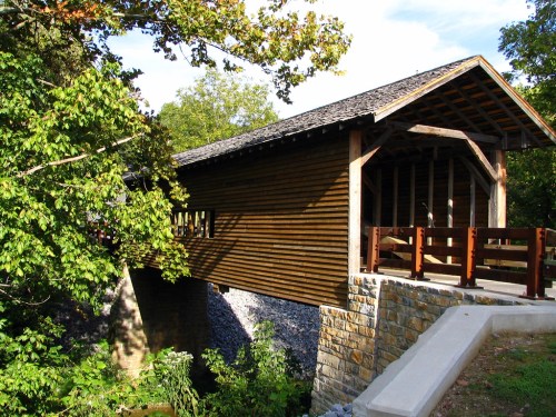 A wooden covered bridge surrounded by trees, spanning over a small stream with a stone foundation.