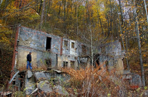 A person stands on rocky ground near the ruins of a dilapidated building, surrounded by autumn foliage and trees.