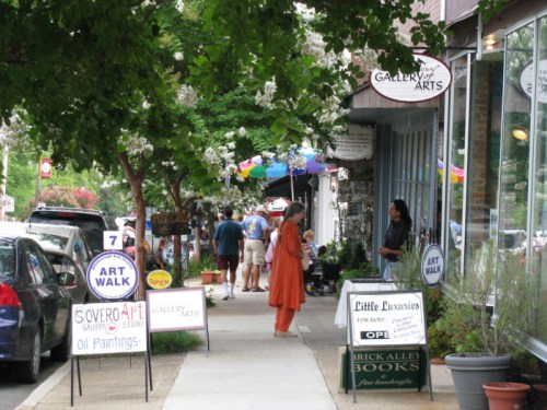 A vibrant street scene with people walking, art displays, and shops during an art walk event. Colorful umbrellas overhead.