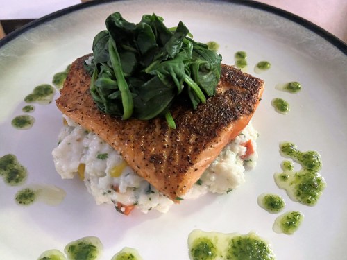A beautifully plated salmon fillet topped with fresh spinach, served on a bed of creamy rice with green sauce drizzled around.