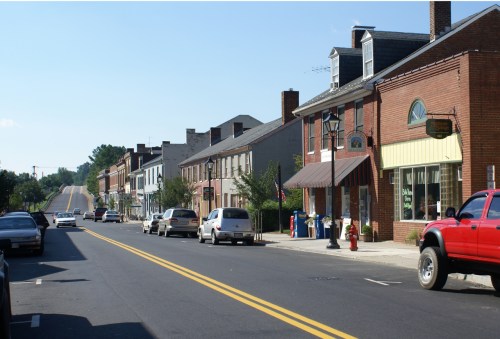 A quiet street lined with brick buildings, shops, and parked cars under a clear blue sky.