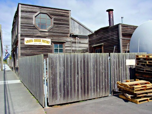 A rustic wooden building with a sign reading "Lovelan Cheese Factory," surrounded by a wooden fence and pallets.