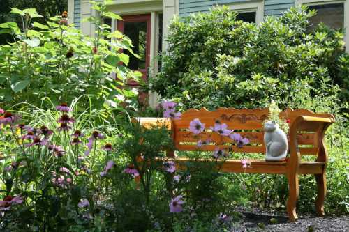 A wooden bench surrounded by colorful flowers and greenery, with a decorative cat statue sitting on it.