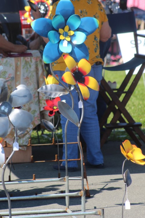 Colorful metal flowers in various designs displayed at an outdoor market, with a person in the background.