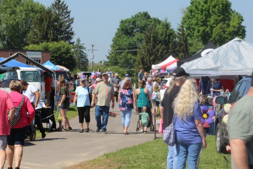 A bustling outdoor market scene with people browsing stalls and enjoying a sunny day. Colorful tents line the path.