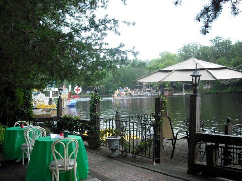A serene riverside scene with tables, a gazebo, and colorful boats in the background surrounded by lush greenery.