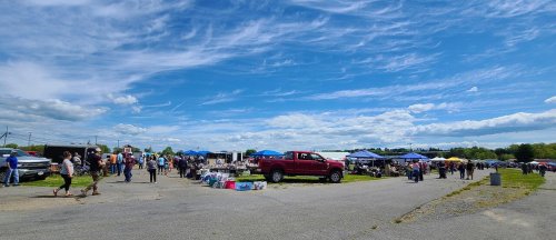 A bustling outdoor market scene with people browsing stalls under a blue sky with scattered clouds.