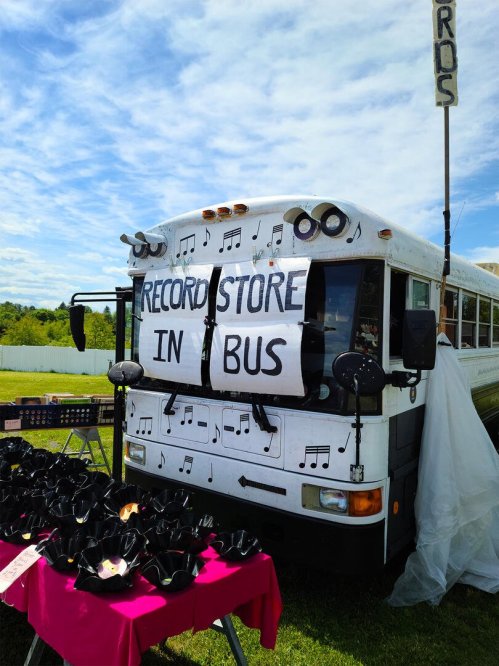 A converted bus with a sign reading "Record Store in Bus," surrounded by vinyl records and black hats for sale.