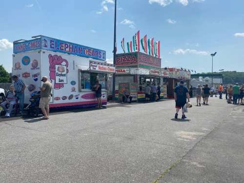 Food trucks lined up at an outdoor event, offering pizza and elephant ears, with people milling about under a clear sky.