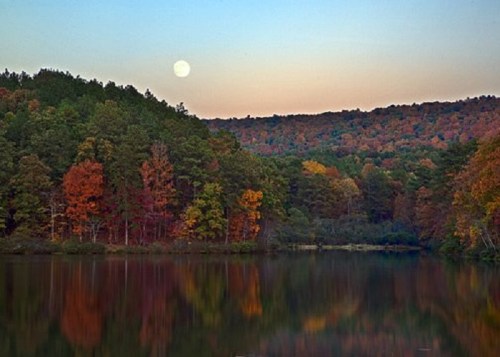 A serene lake reflects colorful autumn trees and a full moon against a twilight sky.