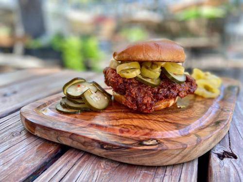 A crispy fried chicken sandwich topped with pickles, served on a wooden platter with additional pickles on the side.
