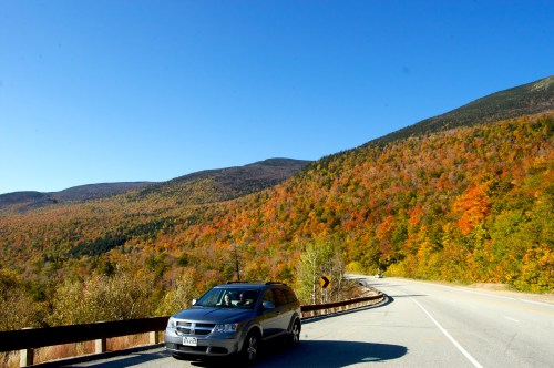 A car parked on a winding road with vibrant autumn foliage and mountains in the background under a clear blue sky.