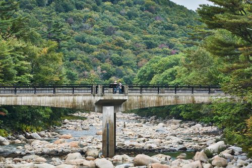 A stone bridge spans a rocky riverbed, surrounded by lush green hills and trees in a serene natural setting.