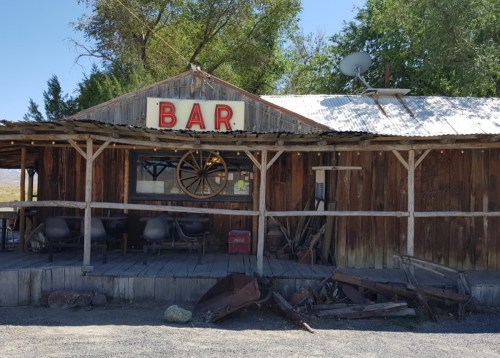 A rustic wooden bar with a sign reading "BAR," surrounded by trees and old furniture outside.