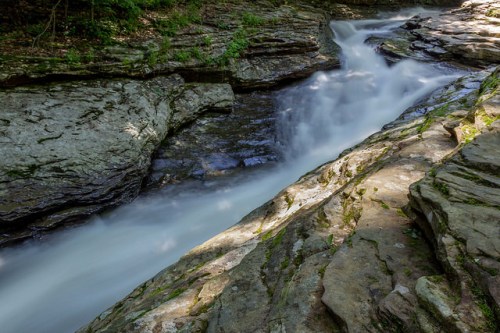 A serene stream flows over smooth rocks, surrounded by lush greenery in a tranquil natural setting.