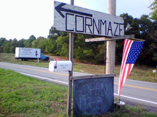 A wooden sign points left to a corn maze, with an American flag nearby and a truck parked on the road. Open 12-8.